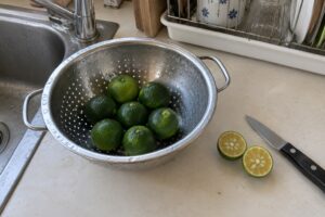 Petits agrumes verts dans une passoire de cuisine, avec un fruit coupé et un couteau près de l’évier.